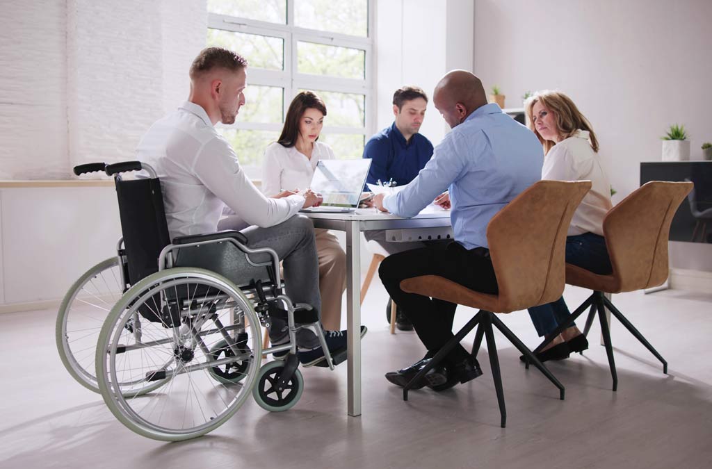 Image of 5 people around a meeting table, one person is in a wheelchair. Representing our adapted property partnerships.