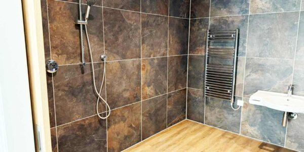 Wheelchair accessible wet-room with modern chrome fittings including a roll under sink. Contemporary natural stone wall tiles and wood effect wet-room flooring.