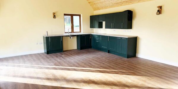 Bright open-plan wheelchair-accessible kitchen diner living room with double height vaulted ceiling. Modern interior with pale lemon coloured walls and a matt black fitted kitchen with roll under sink and pull-down cabinets.